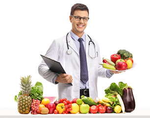 Canvas Print - Doctor holding a clipboard and a plate behind a table with fruit and vegetables