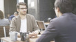 © diproduction - Two friends are smiling talking having a conversation at table of the cafe outdoors. A man dressed in a jacket wearing eyeglasses listening to his friend talking