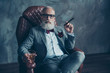 © deagreez - Attractive, old man in spectacles, hold cigarette,  glass with brandy, in tux with red bow-tie and pocket square, sit in leather chair over gray background, looking to the camera, shares, stock, money