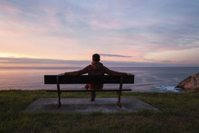 Full Length Of Man Looking At Sea While Sitting On Bench Against Cloudy Sky During Sunset