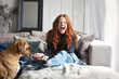 Hübsche rothaarige Frau sitzt lachend auf einer Couch während ein Hund an ihrer Hand schnüffelt
