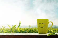 Happiness And Relaxation Concept. A Cup Of Hot Tea With Smiling Face On Table In Front Of Green Tea Plantation Farm, Mountain With Mist As Background