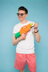 Play as much as possible while you are young! Smiling handsome teenage boy playing with watering spray gun over isolated pale-blue background.