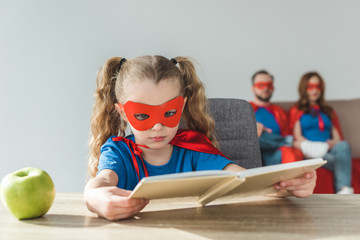 Wall Mural - girl in superhero costume reading book while super parents sitting behind