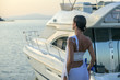 Young beautiful brunette woman relaxing at dock near boat, sunset on summer day. Woman in fashionable clothes stands near luxury yacht, rear view. Luxury life concept.