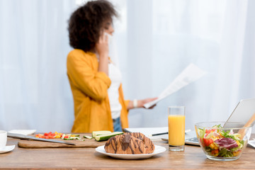 Wall Mural - young woman talking by phone and reading documents with food on table on foreground