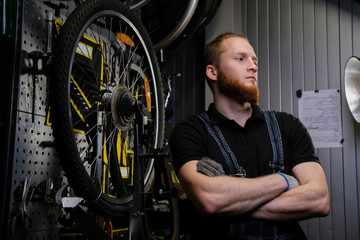 Portrait of a handsome redhead male with beard and haircut wearing jeans coverall, standing near bicycle wheel in a workshop against wall tools.