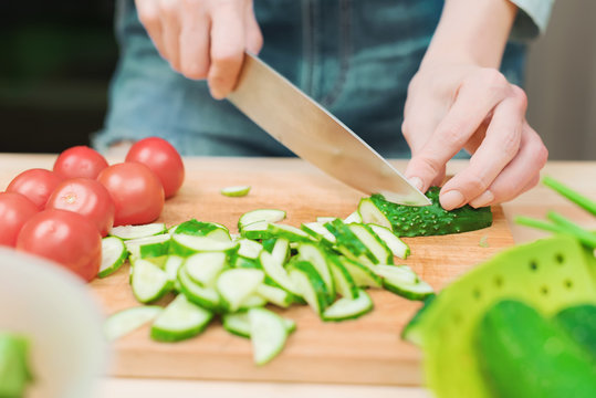 close-up of female hands cut into fresh cut cucumbers on a wooden cutting board next to pink tomatoe