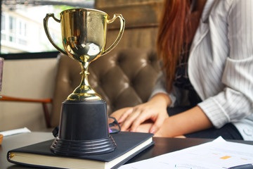 Wall Mural - trophy on working table which have document and pen and black book and bakcground working women