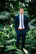© Serhii - Serious portrait handsome groom in a wedding suit and tie is standing on the background of greenery. Man stands in the Botanical Garden full of greenery.