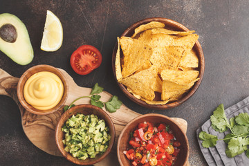 Wall Mural - Mexican food concept. Nachos - yellow corn totopos chips with various sauces in wooden bowls: guacamole, cheese sauce, pico del gallo. Top view, food background