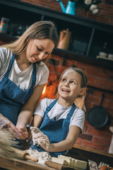 Canvas Print - Cheerful little girl looking at mother and helping with dough preparing on kitchen. 