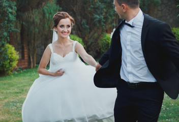 Beautiful wedding couple is running holding hands outdoors in the green park. Bride and groom summer love story. Fun and real emotions.