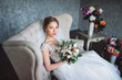 A beautiful bride is sitting in the studio on the couch in a lace dress. Sweet bride in a gray studio. Portrait of the bride.