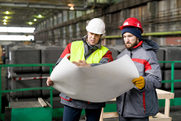 Wall Mural - Portrait of two engineers wearing hardhats holding blueprints and discussing construction in modern industrial workshop, copy space