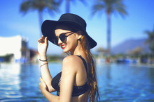 Young Beautiful Girl In Black Fashion Hat Smile, Velvet Skin, Red Lips, Black Swimsuit Posing In The Pool In Blue Water, Stylish Sunglasses, Glamor, Outdoor Portrait, Close Up, Beackground Palms.