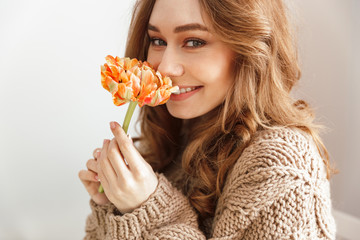 Wall Mural - Photo of lovely woman in sweater sitting at table in cafe looking on camera with smile, and smelling flower