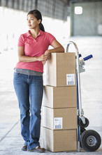 Black Female Standing Next To Hand Truck In Front Of Loading Dock Doors Inside Of A New Warehouse.