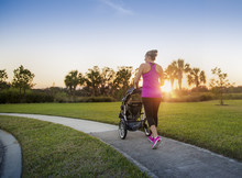 Beautiful, Fit Women Walking And Jogging Outdoors Along A Paved Sidewalk In A Park Pushing A Stroller At Sunset