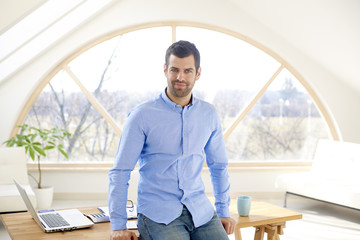 Portrait of young casual businessman looking thoughtful while standing at office desk and smiling. 