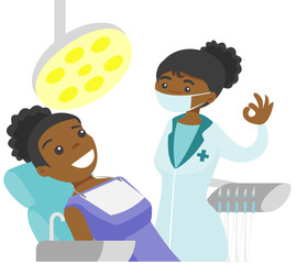 Wall Mural - Young african-american dentist standing next to the dental chair with a patient and showing ok sign in the dental clinic