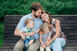 © beatleoff - young laughing family of four sitting on wooden bench. Parents with cute kids.
