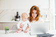 © Anton - Young beautiful business woman sitting at the table and working on laptop while holding her cute little baby isolated.