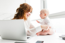 Portrait Of Beautiful Smiling Mother Sitting And Happily Looking At Her Cute Little Baby While Working On Laptop Isolated
