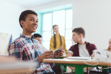 Low angle view of african american high school student holding pencil in hand and classmates sitting behind
