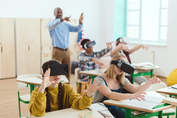 High school teenage students using virtual reality headsets and gesturing teacher standing behind in classroom