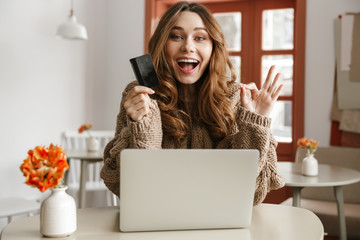 Canvas Print - Photo of pleased brunette woman 20s in sweater doing online shopping with credit card and laptop, while resting in cafe