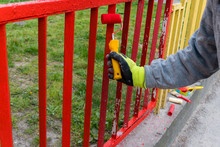 A Worker With Yellow Gloves And Paint Roller Painting A Red Fence On The Background Green Grass