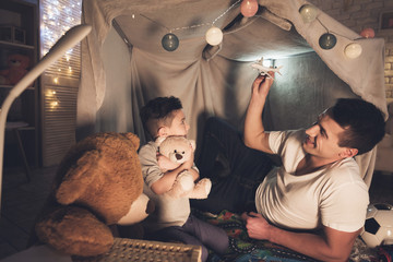 Father and son are playing with toy airplane and cars at night at home.