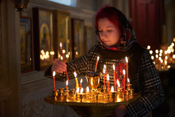 Wall Mural - Praying young woman with candle near pedestal with many other candles in church by her hand to show her faith and esteem to God