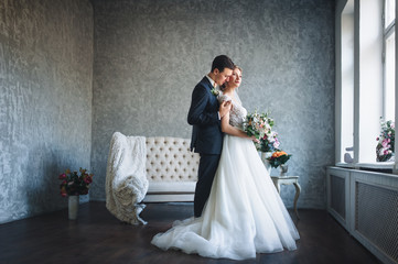 A stylish bridegroom tenderly embraces the bride in a lace dress. The newlyweds embrace in a gray studio with a large sofa and flowers.