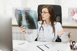 © ViDi Studio - Female doctor sitting at desk, working on computer, holding X-ray of lungs, fluorography, roentgen in light office in hospital. Woman in medical gown, stethoscope in consulting room. Medicine concept.