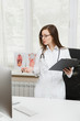 © ViDi Studio - Young female doctor standing at desk, working with clipboard and medical documents in light office in hospital. Woman in medical gown, stethoscope in consulting room. Healthcare, medicine concept.