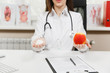 © ViDi Studio - Close up cropped female doctor sitting at desk, holding bottle with white pills in palm, red apple in light office in hospital. Woman in medical gown, stethoscope in consulting room. Medicine concept.
