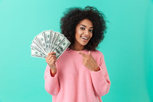 Portrait Of Excited Woman 20s With Afro Hairstyle Pointing Finger On Fan Of Money Dollar Bills, Isolated Over Blue Background