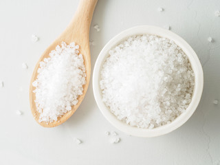 White bowl with large sea salt, wooden spoon,  scattered salt crystals on gray stone table. Top view, close up