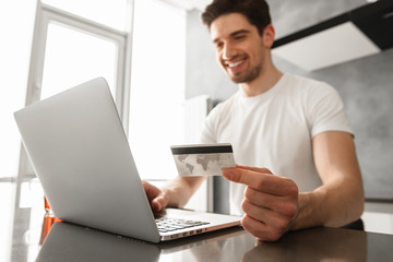 Wall Mural - Photo of successful man 30s in casual clothing holding credit card, and doing payment transaction on laptop in modern apartment