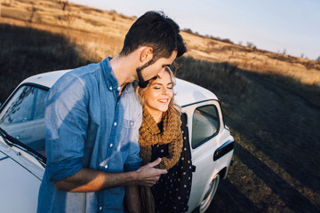 Wall Mural - Young beautiful couple hugging each other, sitting on a small white car in beautiful evening light. Stylish guy with a beard and blond girl laughing