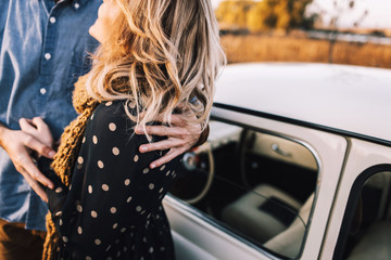 Wall Mural - Young beautiful couple hugging each other, sitting on a small white car in beautiful evening light. Stylish guy with a beard and blond girl laughing