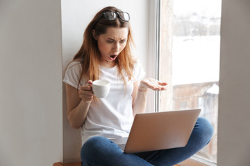 Poster - Shocked woman in t-shirt and eyeglasses sitting on windowsill