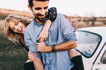 Wall Mural - Young beautiful couple hugging each other, sitting on a small white car in beautiful evening light. Stylish guy with a beard and blond girl laughing