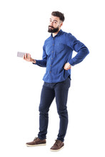 Uncertain Confused Bearded Business Man Holding Mobile Phone And Looking At Camera. Full Body Isolated On White Studio Background. 