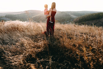 beautiful young mother in a long red dress easy walks with her young son. Summer, sunset, high yellow grass, mountains. He is on the hands, holding the hand