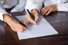 Closeup Of Two Business Women Signing Contract Together. They Are Holding Pens And Going To Write On Blank Paper Sheet. Contract Concept. Front View.