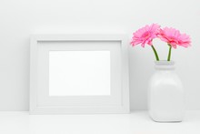 Mock Up White Frame And Pink Daisy Flowers In Vase On A Shelf Or Desk. White Color Scheme. Landscape Frame Orientation.