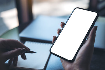 Mockup image of a woman's hand holding black mobile phone with blank white desktop screen with laptop and notebook on table